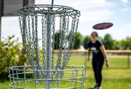A golf disc hole in a park on a sunny day with a disc in the air after being tossed by a player