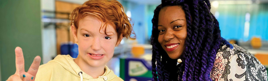 A student and an educator posing for a picture. The student makes a peace sign with one hand and has blue slime in the other hand.