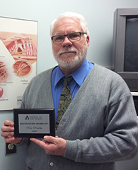 Reg Dunphy wearing a grey sweater with a tie and blue shirt holding a plaque