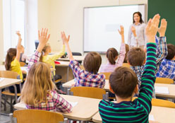 Young students in a classroom with their hands raised waiting to be called on by the teacher at the head at the classroom.