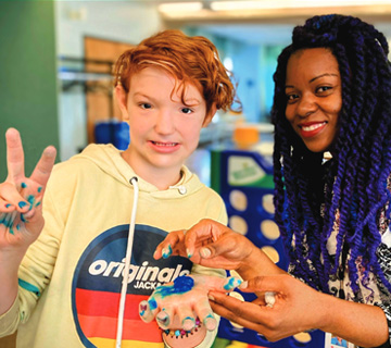 A student and an educator posing for a picture. The student makes a peace sign with one hand and has blue slime in the other hand.