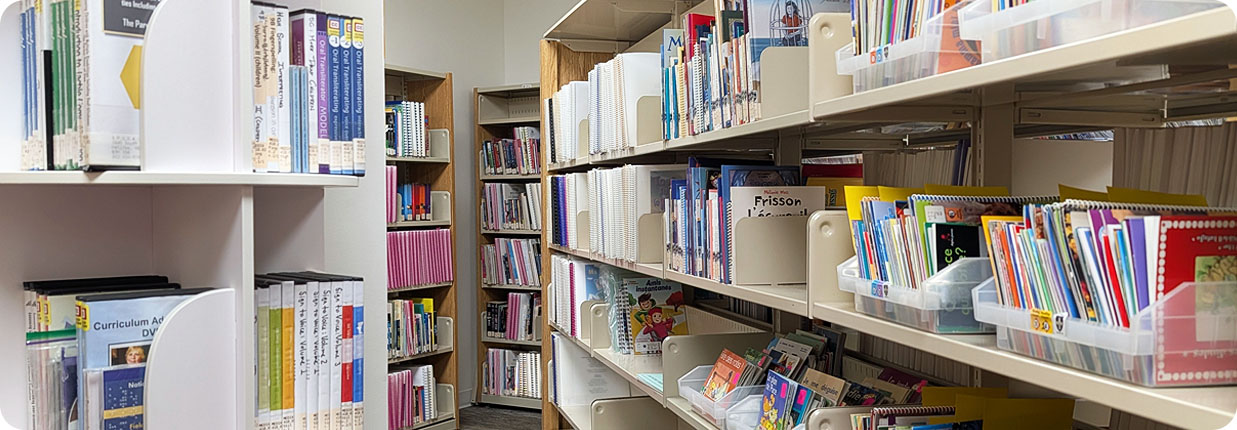 Shelves in a library filled with books and educational materials, including binders, textbooks, and organized bins of resources.
