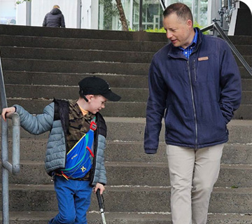 A child using a white cane walks down outdoor concrete steps while holding a metal handrail. An adult walks beside them, watching attentively and offering support.
