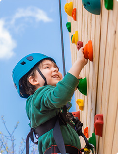 Child wearing a blue helmet and harness climbing an outdoor wooden rock‑climbing wall with colorful handholds