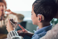 A young boy with a hearing aid holds a smartphone, smiling, as an adult gestures nearby. The scene conveys communication and connection in a warm, supportive environment.