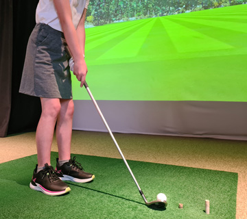 Person holding a golf club and preparing to swing at a ball on an indoor golf simulator turf, with a projected fairway scene in the background.