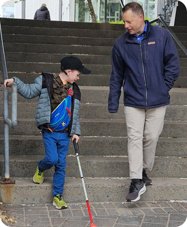 A child using a white cane walks down outdoor concrete steps while holding a metal handrail. An adult walks beside them, watching attentively and offering support.