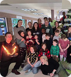 A large group of children and adults gather around a Christmas tree in a school. Many smile and pose for the photo, while some children stand, kneel, or sit at the front. One performer kneels holding a flaming prop.