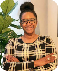 A smiling woman stands indoors with her arms crossed, wearing glasses and a black-and-cream plaid top. Her hair is styled in a high bun, and she wears small earrings and a ring. A leafy green plant is visible behind her against a light-coloured wall.