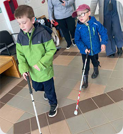 Two children walk indoors along a tiled hallway using white canes for orientation and mobility practice. One child wears a green jacket, the other wears a blue jacket, glasses and a cap, following alongside.