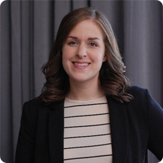 Brigitte Hanna, les cheveux bruns mi-longs, sourit doucement, vêtue d'un blazer noir sur une chemise rayée, devant un rideau gris en arrière-plan.