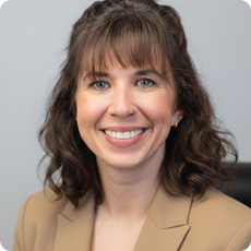Person with shoulder‑length curly hair wearing a tan blazer, seated against a plain background.