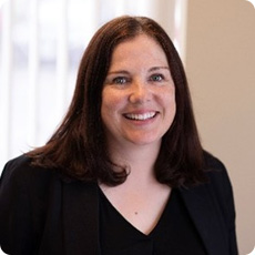 Sarah Coughlan with shoulder-length dark hair, wearing a black top and blazer, smiles warmly in a softly lit room. The background is softly blurred.